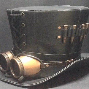 Puede incluir: Un sombrero de copa de cuero negro con una banda de cuero marr&oacute;n y gafas. El sombrero tiene un detalle de encaje en el lateral y una correa de cuero con una fila de balas de metal en la parte posterior.