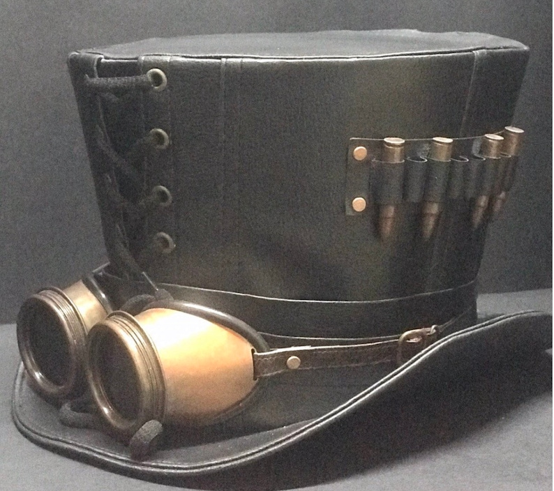 Puede incluir: Un sombrero de copa de cuero negro con una banda de cuero marr&oacute;n y gafas. El sombrero tiene un detalle de encaje en el lateral y una correa de cuero con una fila de balas de metal en la parte posterior.