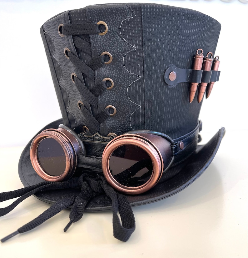 May include: A black top hat with a leather band and a lace-up detail. The hat has a pair of goggles attached to the front, with copper-colored frames and dark lenses. There are also several small, copper-colored bullet casings attached to the side of the hat.