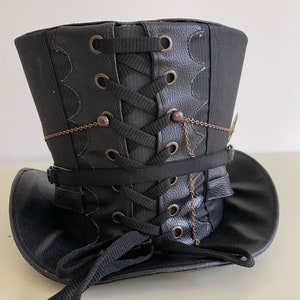 Pu&ograve; includere: Cappello a cilindro nero con design steampunk. Il cappello presenta una texture in similpelle, lacci decorativi e dettagli a catena color rame. La tesa &egrave; curva e il cappello ha una forma strutturata e verticale.