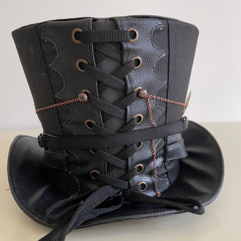 Pu&ograve; includere: Cappello a cilindro nero con design steampunk. Il cappello presenta una texture in similpelle, lacci decorativi e dettagli a catena color rame. La tesa &egrave; curva e il cappello ha una forma strutturata e verticale.