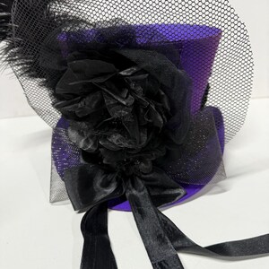 Pu&ograve; includere: Un mini cappello a cilindro viola con dettagli neri. Il cappello presenta un velo a rete nero, un grande fiore nero e un fiocco nero con lunghi nastri. Una piuma nera &egrave; attaccata al lato del cappello.