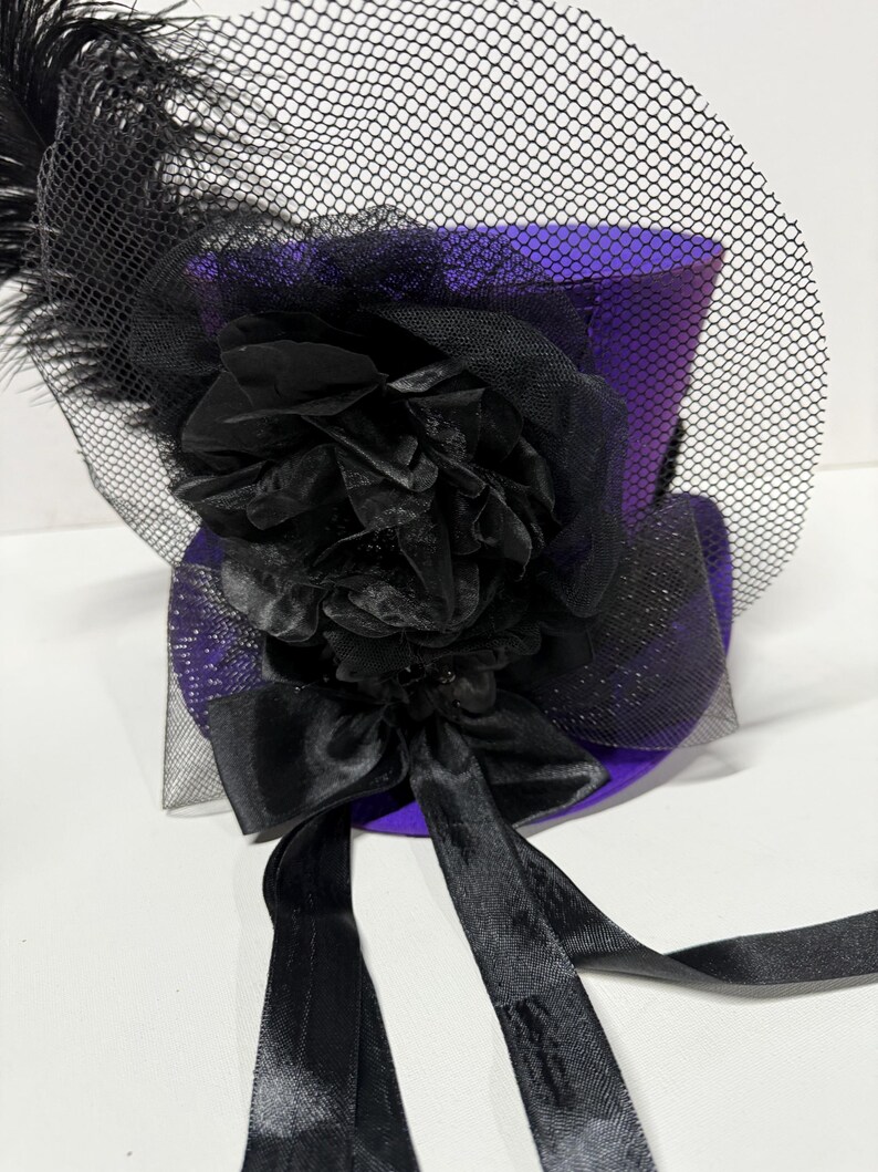 Pu&ograve; includere: Un mini cappello a cilindro viola con dettagli neri. Il cappello presenta un velo a rete nero, un grande fiore nero e un fiocco nero con lunghi nastri. Una piuma nera &egrave; attaccata al lato del cappello.