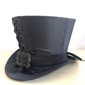 Op de afbeelding: Zwarte hoge hoed met een decoratieve zilveren gesp en zwart lint. De hoed heeft een vetersluiting aan de zijkant en een brede rand. Deze hoed is geschikt voor cosplay, steampunk of kostuumevenementen.