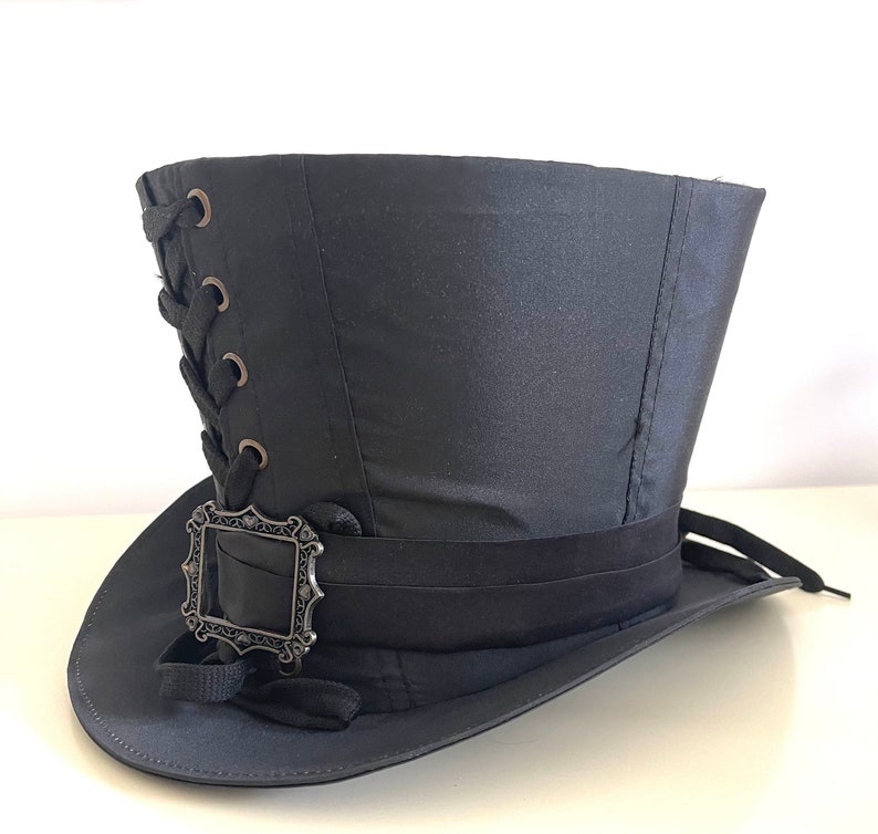 Op de afbeelding: Zwarte hoge hoed met een decoratieve zilveren gesp en zwart lint. De hoed heeft een vetersluiting aan de zijkant en een brede rand. Deze hoed is geschikt voor cosplay, steampunk of kostuumevenementen.