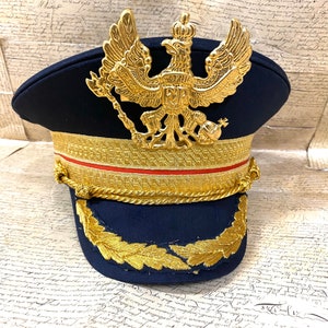 May include: A navy blue military style hat with a gold braid trim and a gold eagle emblem on the front. The eagle has a crown on its head and is holding a scepter in its right talon.