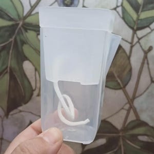 May include: A translucent, rectangular plastic container featuring a removable lid and a white, flexible cord coiled inside. The container has a small, angled spout on one side and a clear, smooth finish, providing a view of the internal cord.