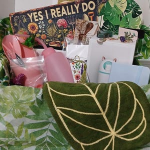 May include: A gift box filled with gardening-themed items. Includes a green leaf-shaped pillow, a sign that reads "YES I REALLY DO", pink watering cans, and other decorative items. The box is lined with green leaf-patterned tissue paper.