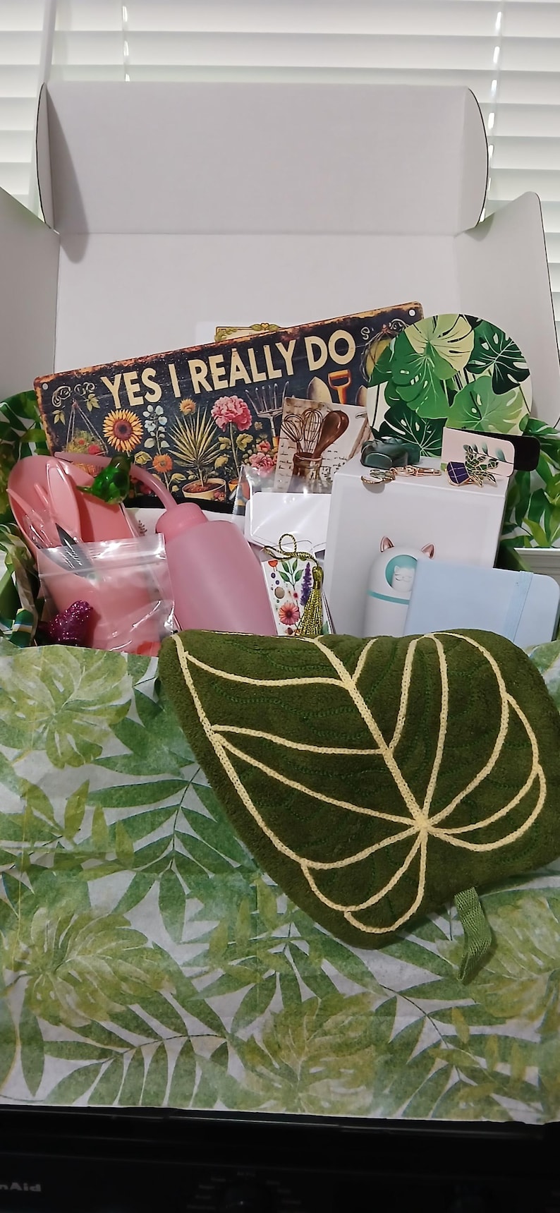 May include: A gift box filled with gardening-themed items. Includes a green leaf-shaped pillow, a sign that reads "YES I REALLY DO", pink watering cans, and other decorative items. The box is lined with green leaf-patterned tissue paper.