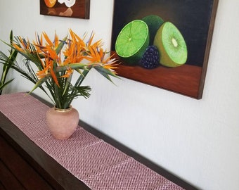 Op de afbeelding: Twee ingelijste schilderijen van fruit hangen aan een witte muur. Eén toont een sinaasappel met geschilde delen, de andere een limoen, kiwi en bramen. Een bordeauxrood en wit patroon tafelloper met franjes ligt op een donkere houten kast. Een vaas met oranje bloemen staat op de kast.