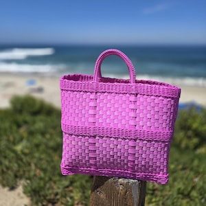 May include: A bright pink tote bag featuring a woven plastic construction with a rectangular body and two integrated top handles. The bag displays a consistent grid-like pattern throughout its surface, showcasing a vibrant solid colour finish.