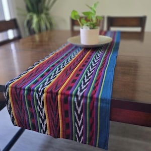 May include: A rectangular table runner featuring a vibrant, multi-colored ikat pattern with vertical stripes in red, yellow, green, blue, and black. The fabric displays a repeating chevron-like design throughout, finished with solid blue borders.