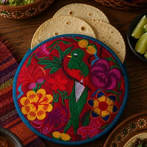 Puede incluir: Un calentador de tortillas redondo de tela roja con bordados multicolores de un quetzal verde rodeado de flores en tonos amarillos, morados y rosas, rematado con un borde de tela azul sólido a lo largo de todo el contorno circular.