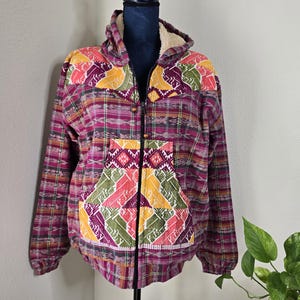May include: A purple, green, yellow and orange patterned hooded jacket with a zip closure. The jacket has a fleece lining and a large embroidered patch on the front.