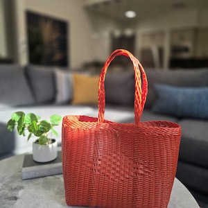 May include: A woven red tote bag with two matching handles. The bag is made of a textured material and sits on a round, light-colored table. The background is blurred, showing a living room setting.
