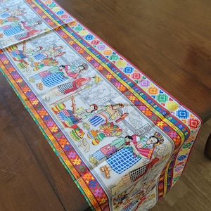 May include: A white table runner with a colorful border featuring a repeating pattern of hearts and geometric shapes. The center of the runner features a colorful illustration of people in traditional clothing.