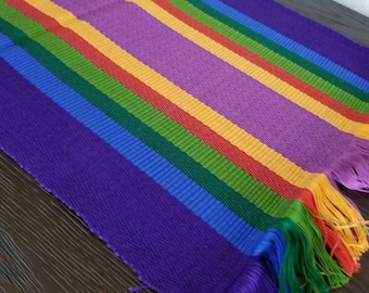 Op de afbeelding: Een kleurrijke tafelrunner met een regenboogstrepenpatroon en paarse franjes. De runner is gemaakt van geweven stof en heeft een textuur.