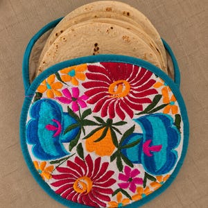 Puede incluir: Un calentador de tortillas redondo de tela con borde color verde azulado y una parte superior blanca con bordados florales vibrantes en tonos rojo, azul, naranja y rosa. El calentador contiene una pila de tortillas de harina beige visibles.