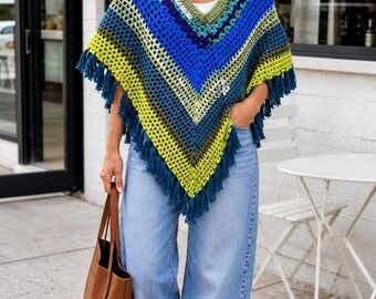 Puede incluir: Un poncho de ganchillo con cuello en V y patrón de chevrón en tonos azul, verde y lima, acabado con flecos azul oscuro a lo largo del dobladillo. La prenda se combina con vaqueros anchos de color azul claro, un bolso de cuero marrón y zuecos.