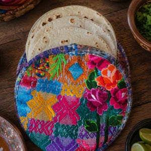 Puede incluir: Un calentador de tortillas redondo de tela con bordados florales y geométricos de colores vibrantes en tonos rosa, naranja, amarillo, azul y verde. El calentador contiene una pila de tortillas de harina redondas de color beige claro.