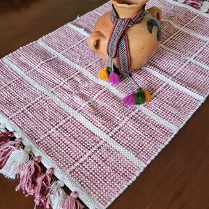May include: A red and white woven table runner with fringe tassels. The runner has a terracotta pot with a black and white patterned ribbon tied around it. The pot has a small handle on each side.
