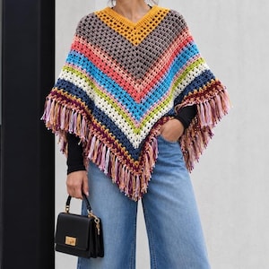 May include: A V-neck crochet poncho featuring a multi-colored chevron pattern in shades of yellow, gray, pink, blue, white, and dark purple. The garment has an open-weave construction and is finished with long, multicolored fringe along the bottom hem.