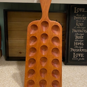 May include: A wooden egg holder with a handle. The holder has 20 indentations for eggs. The wood is a reddish brown color.