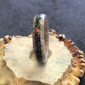 May include: A white antler ring with a thin band of iridescent opal inlay. The ring is displayed on a white antler base.