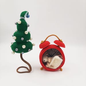May include: A green crocheted Christmas tree with pearl and silver bead accents stands next to a red crocheted alarm clock. The clock face is visible, and a small crocheted animal is nestled inside.