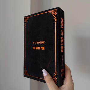May include: Black book with gold-colored accents and text. The book's spine reads "HUNT THE VILLAIN" vertically. The front of the book has text in both English and Cyrillic script. Decorative gold-colored filigree borders the edges.