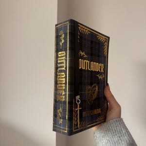 Puede incluir: Un libro de tapa dura titulado "Outlander" con un diseño de cuadros negros y grises y letras doradas. El libro tiene detalles florales dorados y un cierre decorativo dorado.