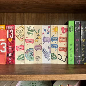 May include: A bookshelf displays a collection of books and decorative bookmarks. The bookmarks are hand-painted with various designs, including a sports jersey, a wing, and speech bubbles with text. The books have titles like "Binding 13" and "Kiss the Villain".