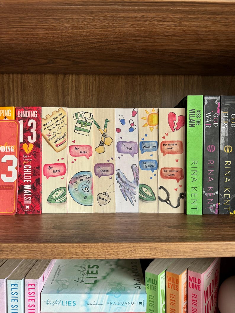 May include: A bookshelf displays a collection of books and decorative bookmarks. The bookmarks are hand-painted with various designs, including a sports jersey, a wing, and speech bubbles with text. The books have titles like "Binding 13" and "Kiss the Villain".