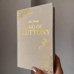 May include: A hardcover book with a beige fabric cover featuring gold foil lettering that reads Ana Huang KING OF GLUTTONY. The cover is decorated with gold foil line art illustrations of roses and a sealed envelope in the top left and bottom right.