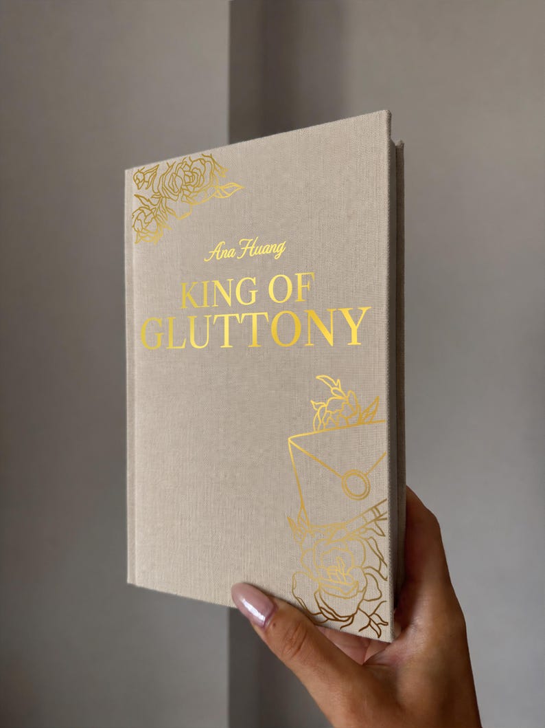 May include: A hardcover book with a beige fabric cover featuring gold foil lettering that reads Ana Huang KING OF GLUTTONY. The cover is decorated with gold foil line art illustrations of roses and a sealed envelope in the top left and bottom right.