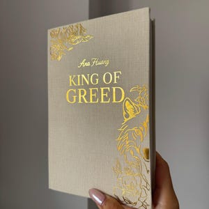 May include: A hardcover book with a beige fabric cover featuring gold foil stamping. The front cover displays the text Ana Huang and KING OF GREED in gold, accompanied by gold foil illustrations of a wolf and floral patterns in the corners.