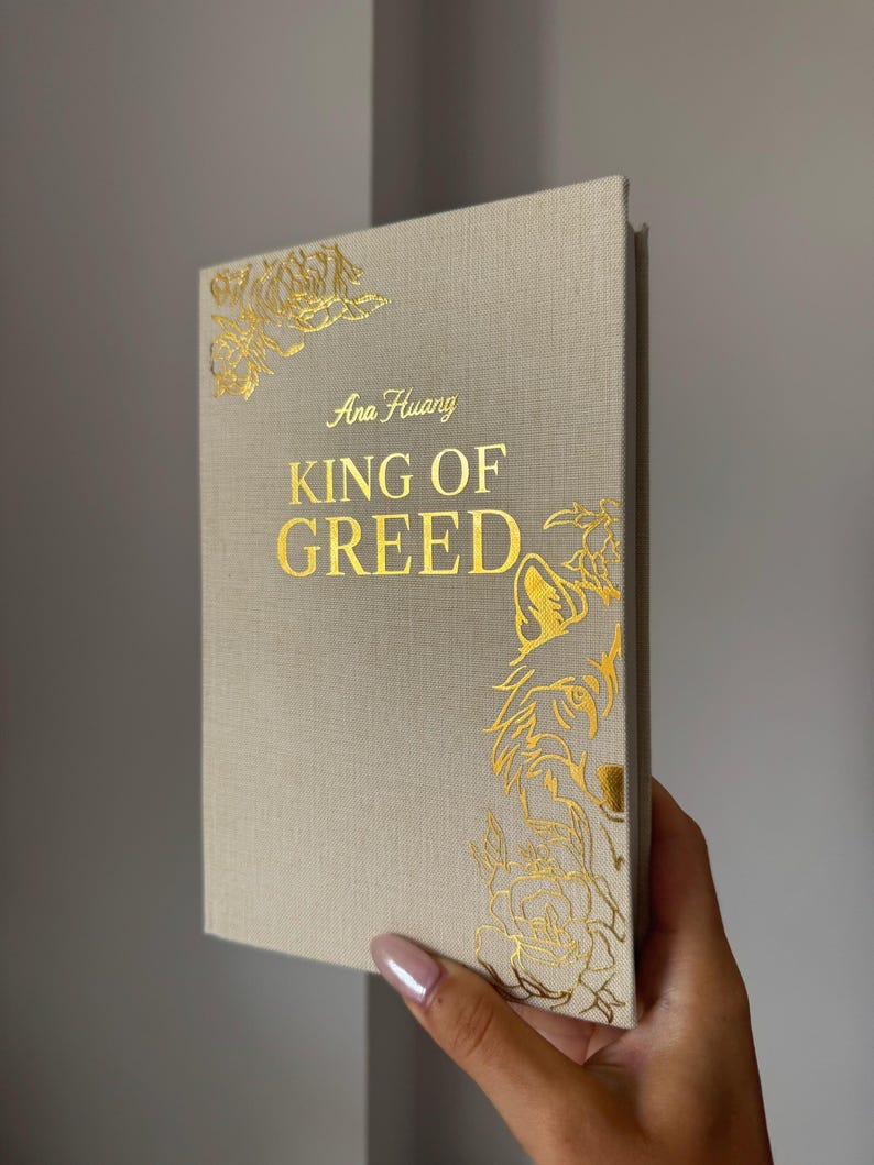May include: A hardcover book with a beige fabric cover featuring gold foil stamping. The front cover displays the text Ana Huang and KING OF GREED in gold, accompanied by gold foil illustrations of a wolf and floral patterns in the corners.