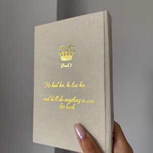 May include: A beige hardcover book with gold foil lettering and a crown graphic on the front cover. The text reads: Kings of Sin, Book 3, He had her, he lost her... and he'll do anything to win her back. The book has a textured fabric cover.