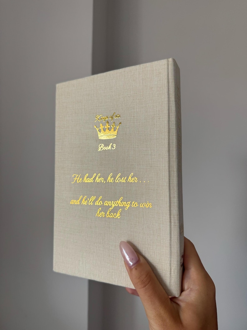 May include: A beige hardcover book with gold foil lettering and a crown graphic on the front cover. The text reads: Kings of Sin, Book 3, He had her, he lost her... and he'll do anything to win her back. The book has a textured fabric cover.