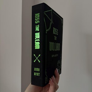 May include: A black book titled "Kiss the Villain" by Rina Kent. The book's spine features the title in green, along with crossed arrows and the author's name. The cover has an ornate design.