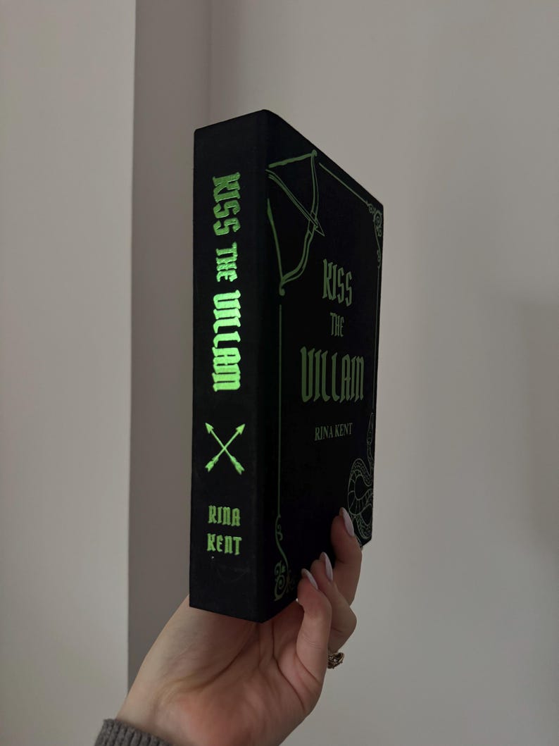 May include: A black book titled "Kiss the Villain" by Rina Kent. The book's spine features the title in green, along with crossed arrows and the author's name. The cover has an ornate design.