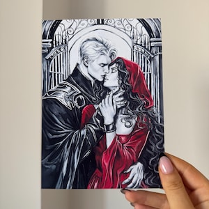 May include: Illustration of a couple embracing, set against a black and white arched gate background. The man has white hair and wears a black coat. The woman wears a red hooded dress. The artwork is held by a hand.