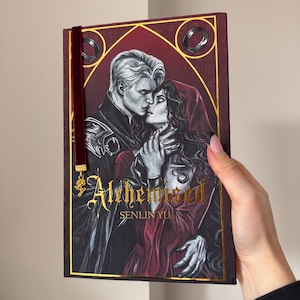 Op de afbeelding: Hardcover boek getiteld "Althemissed" van Senlin Yu. De omslag toont een romantische illustratie van een stel in een gepassioneerde omhelzing, met een donkerrode achtergrond en gouden accenten. Een bordeauxrood fluwelen bladwijzer is bevestigd.