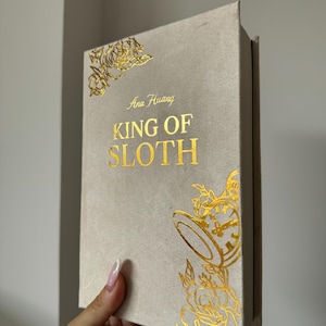 May include: A hardcover book titled King of Sloth by Ana Huang with a light beige fabric cover. The front cover features gold foil lettering and decorative gold foil illustrations of roses and a pocket watch in the corners.
