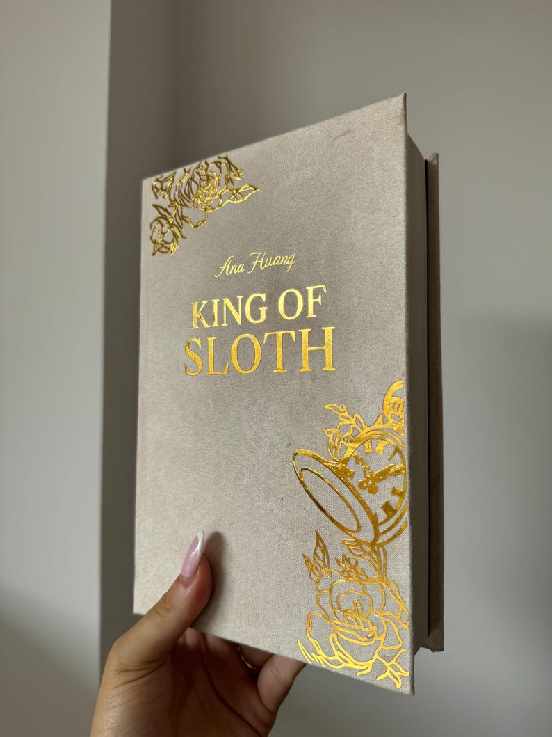 May include: A hardcover book titled King of Sloth by Ana Huang with a light beige fabric cover. The front cover features gold foil lettering and decorative gold foil illustrations of roses and a pocket watch in the corners.