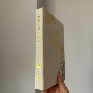 May include: A hardcover book titled King of Greed by Ana Huang with a textured beige fabric cover. The spine and front cover feature gold foil lettering and decorative gold floral illustrations, including a small crown icon on the spine.