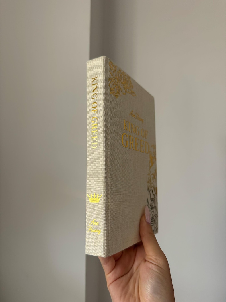 May include: A hardcover book titled King of Greed by Ana Huang with a textured beige fabric cover. The spine and front cover feature gold foil lettering and decorative gold floral illustrations, including a small crown icon on the spine.