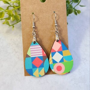 May include: A pair of teardrop-shaped earrings with a geometric pattern in shades of blue, green, pink, yellow, and white. The earrings are made of wood and have silver hooks.