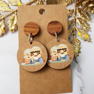 May include: A pair of wooden earrings featuring a cartoon image of an elderly couple holding mugs. The earrings are brown with a wood grain pattern and have silver hooks.