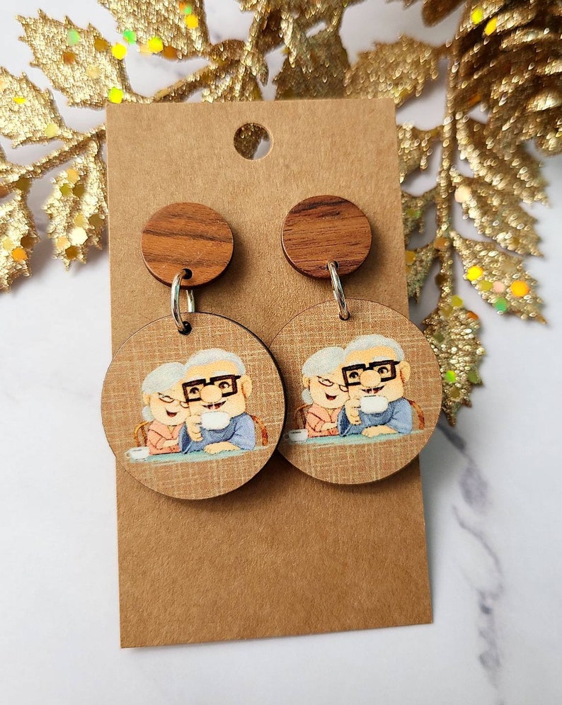 May include: A pair of wooden earrings featuring a cartoon image of an elderly couple holding mugs. The earrings are brown with a wood grain pattern and have silver hooks.